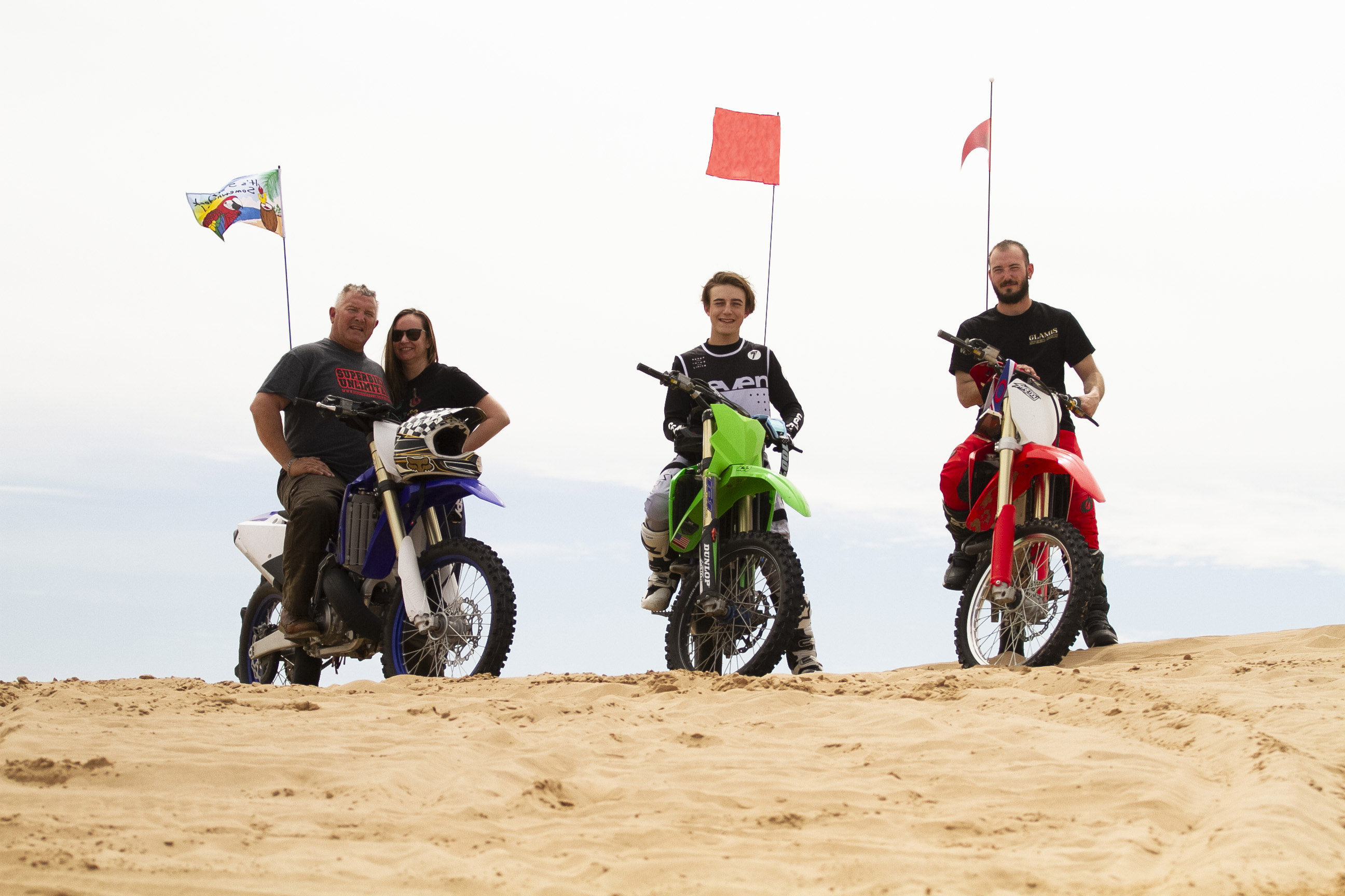 Four people posed on a dune with dirt bikes. 