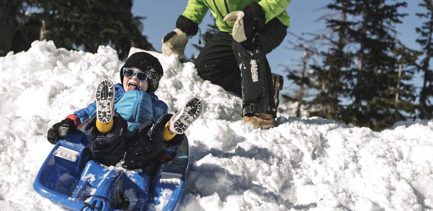 A young boy sleds down snow as his grandfather pushes him.