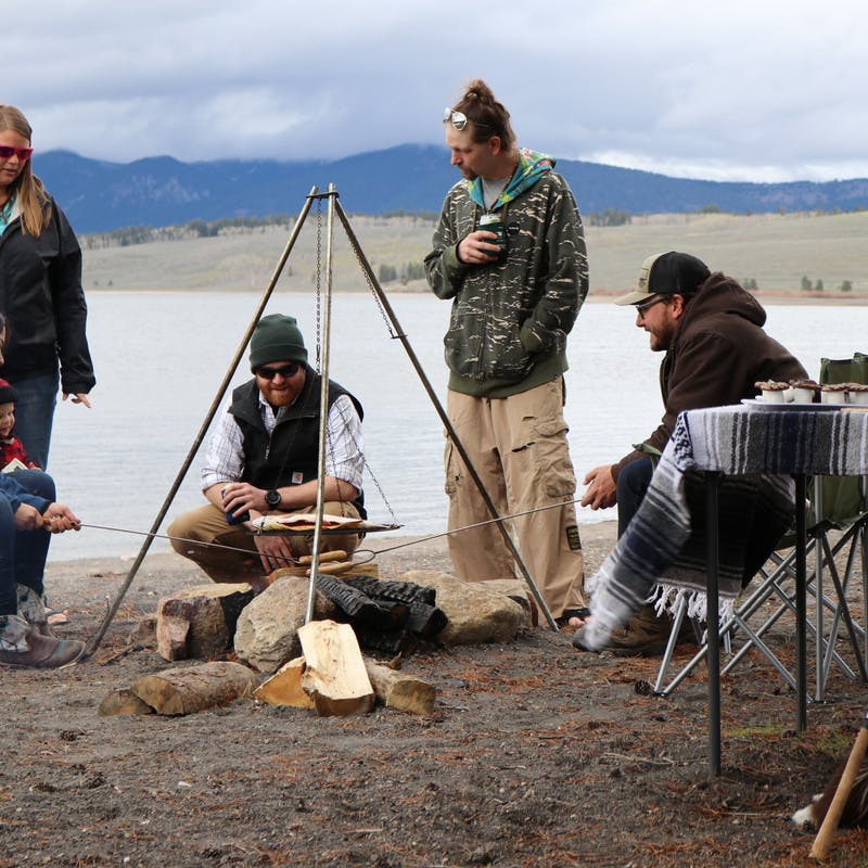 Group of five friends and one child stands around a campfire cooking hot dogs on a stick, with lake in the background.