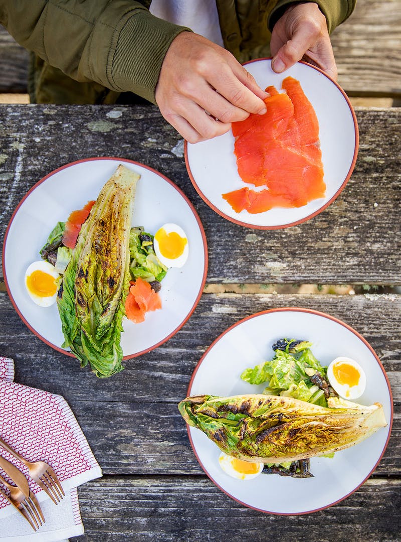 Man holds plate with lox, while preparing his caesar salad dishes with romaine hearts and hard boiled eggs.