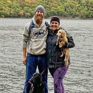 Kelsey and Scott Kiessig pose for the camera with their 2 dogs, while wearing sweatshirts, jackets, and winter hats in front of a body of water and trees.