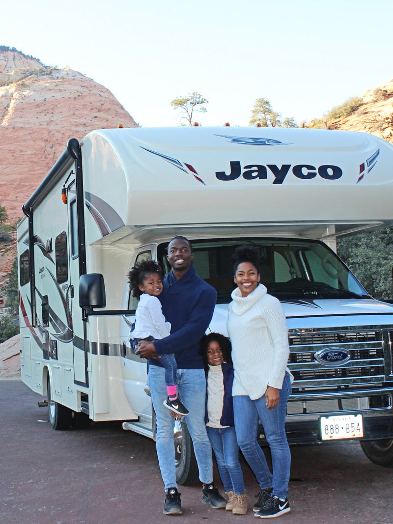 An African American family of 4 stands outside of their Jayco RV that is pulled off on the side of the road with the mountains and trees in the background.