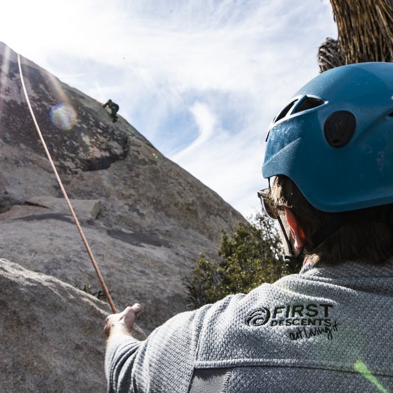 Volunteer wearing a First Descents jacket holds a climber's rope steady.