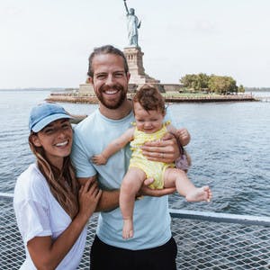 Bryce, Nellie and Avalyn Jurgy, woman and man holding a baby, on a boat, posing in front of the Statue of Liberty.