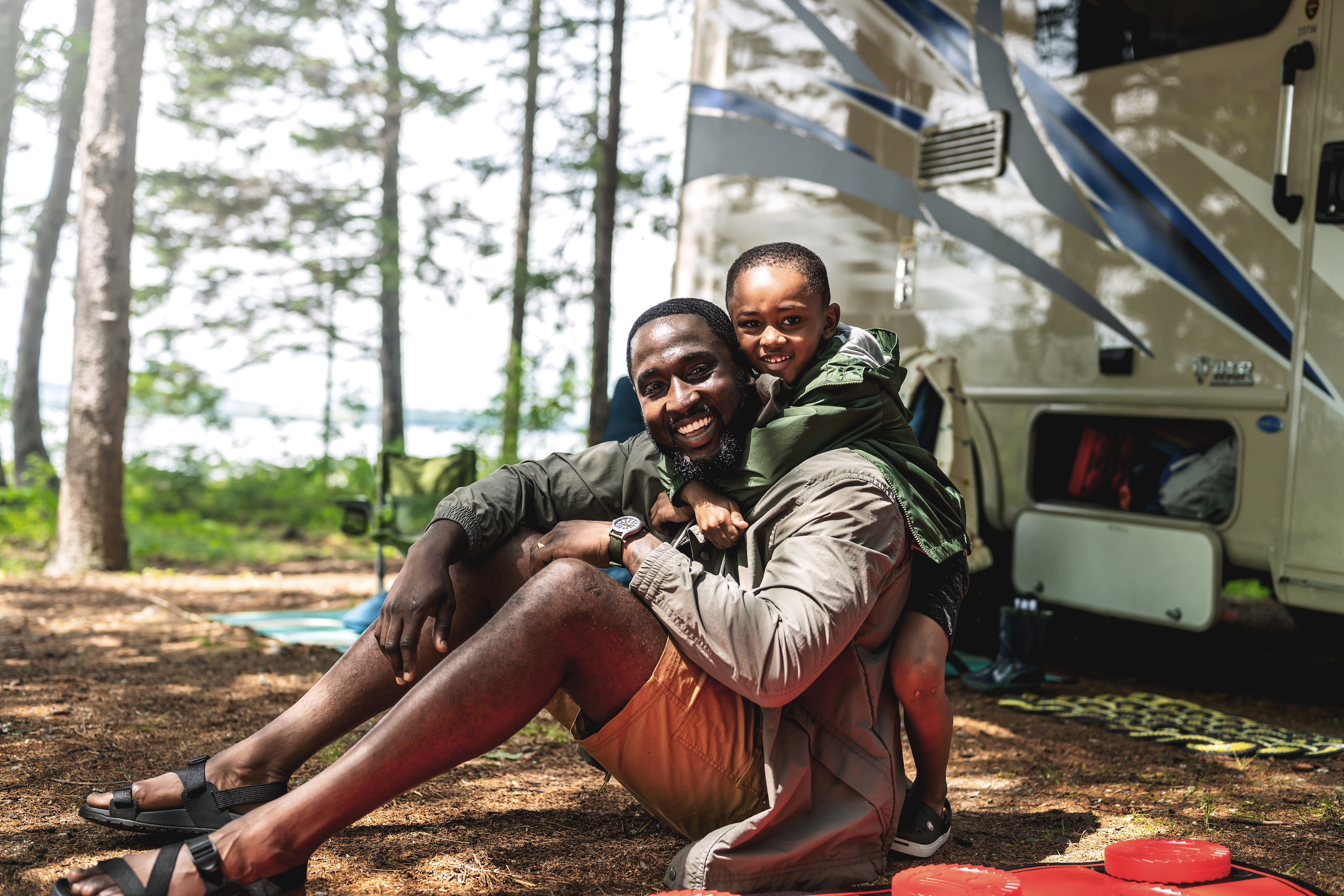 A son hugs his father from behind as they sit near an RV