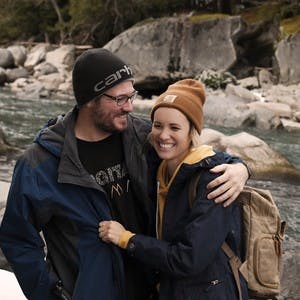 Mel and Tony Flammia, wearing winter clothes, laughing and smiling for a photo with a creek filled with rocks behind them.