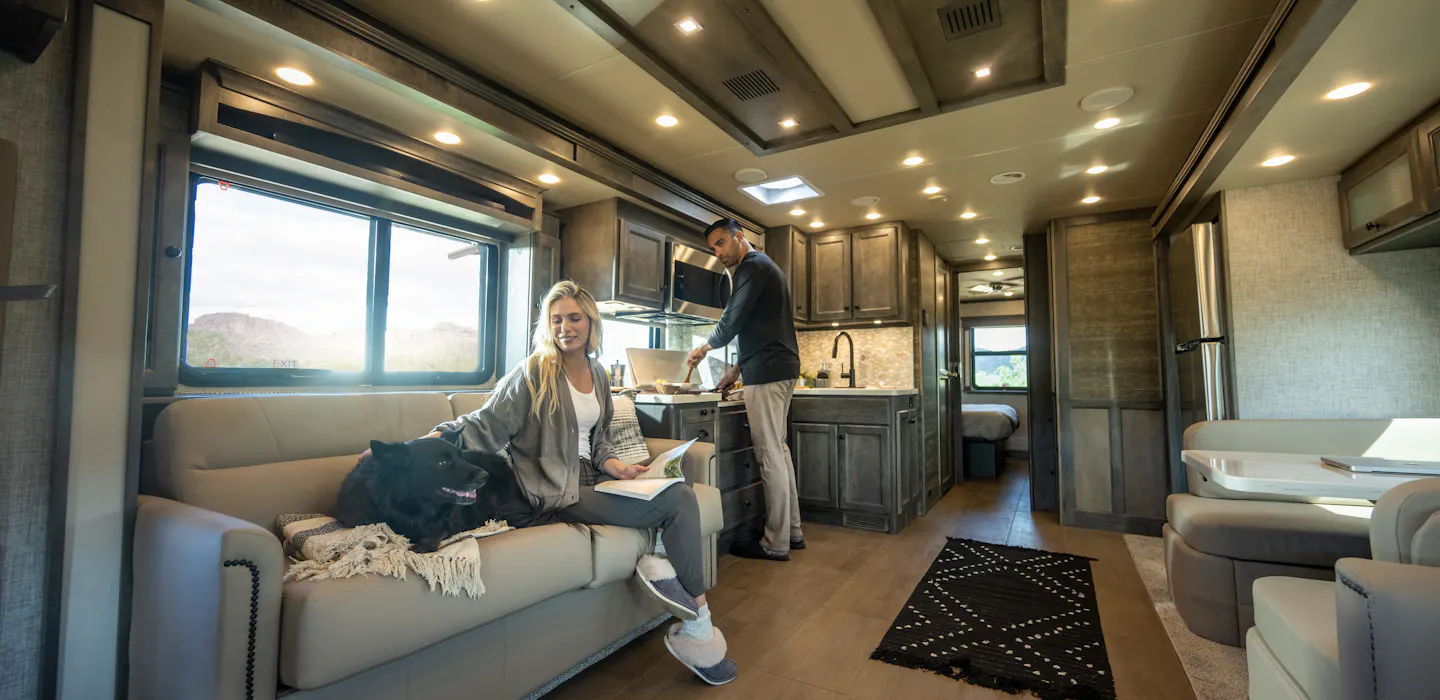 A couple is seen inside a Tiffin Allegro Bay Super C, where one person is cooking while the other is sitting on the couch petting a dog.
