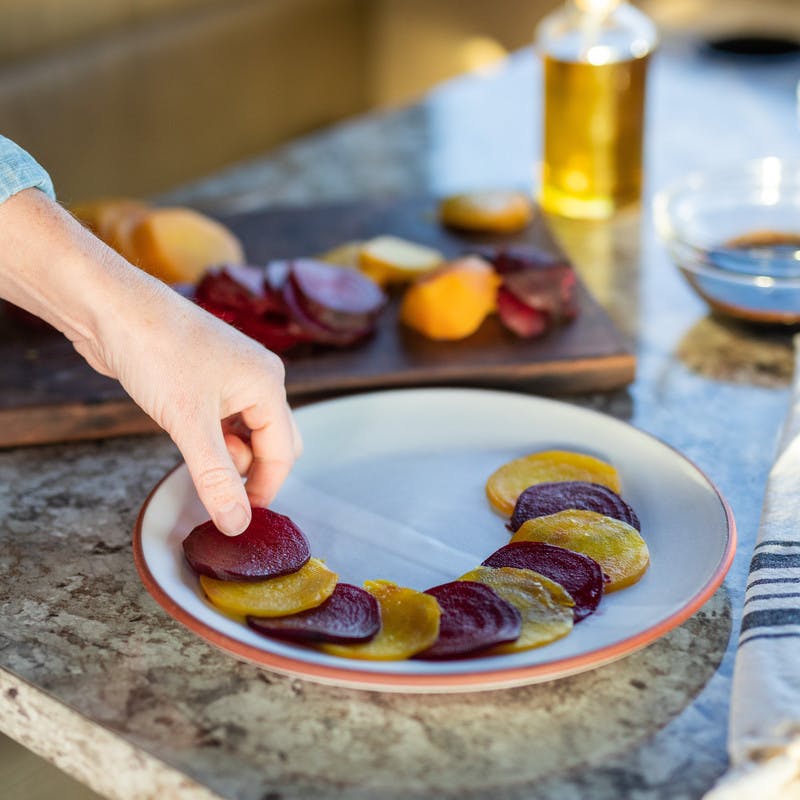 Hand places sliced red and gold beets into a circle on a white plate, with chopped beats and a bowls in the background.