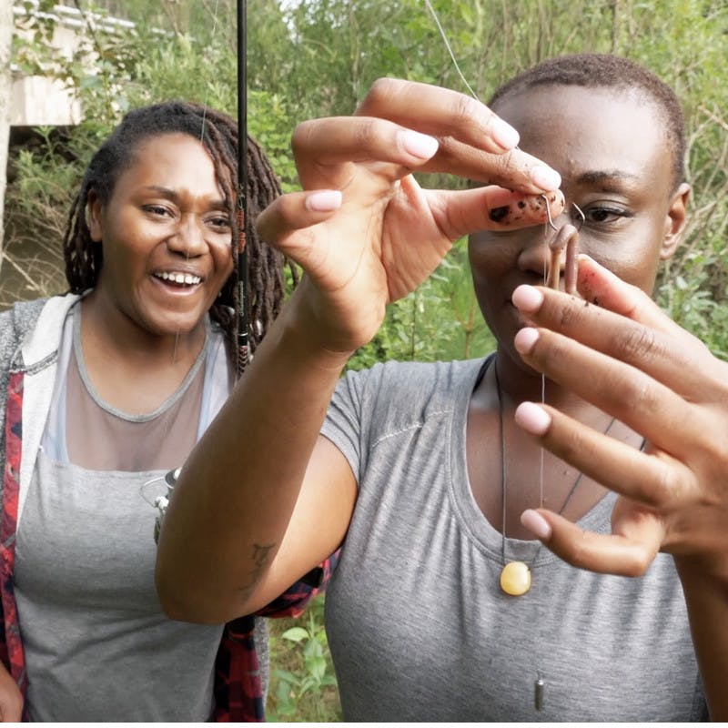 Chanel and Brittany Tate hooking bait onto a fishing line by a river.