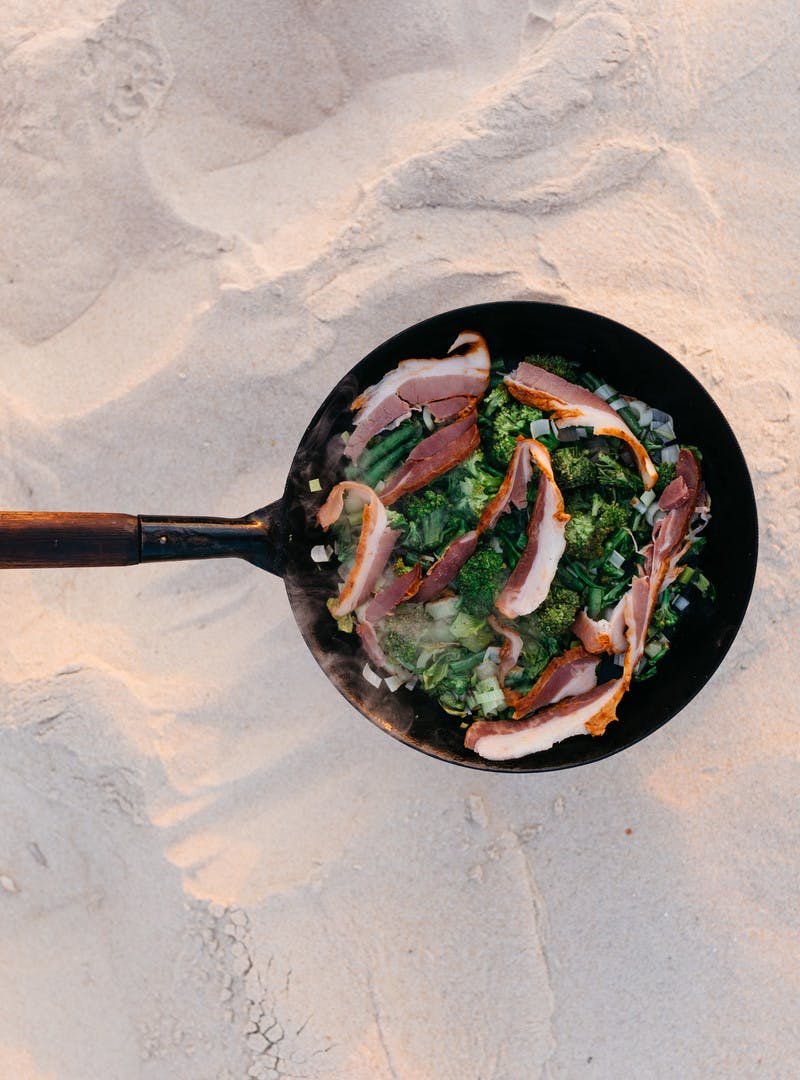 Greens, green beans and leeks cooking in a pan with bacon cooking on top.