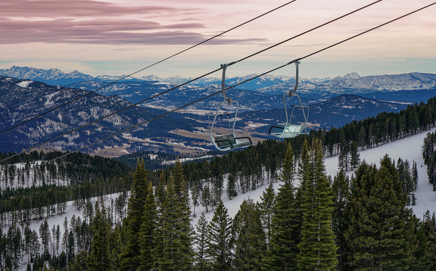 Ski lift chairs hanging above green pines and snowy mountains in the background with pink skies