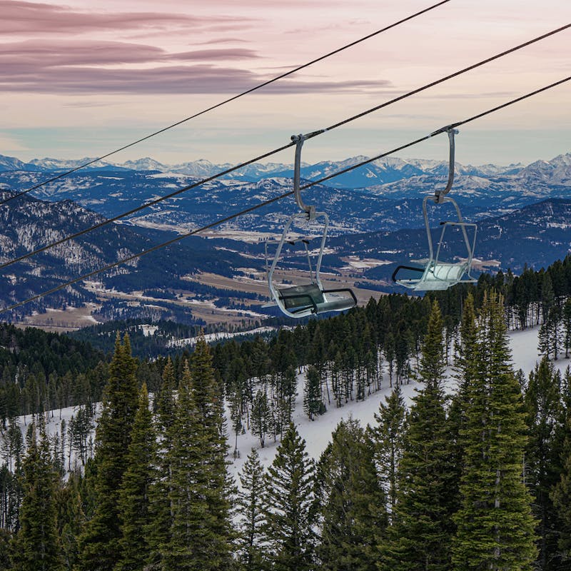 Ski lift chairs hanging above green pines and snowy mountains in the background with pink skies