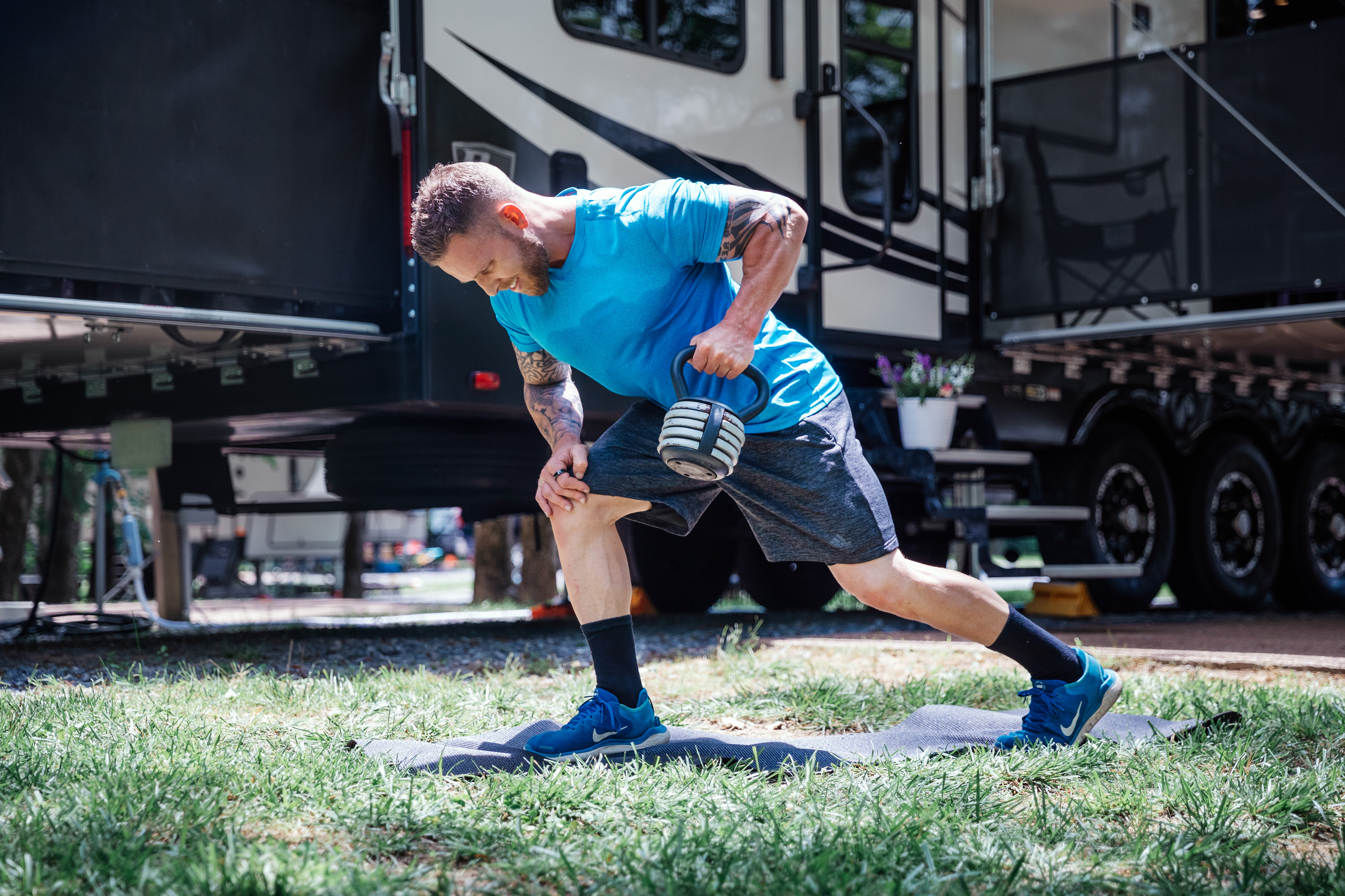 A muscled man doing one-armed rows with a kettlebell outside an RV.