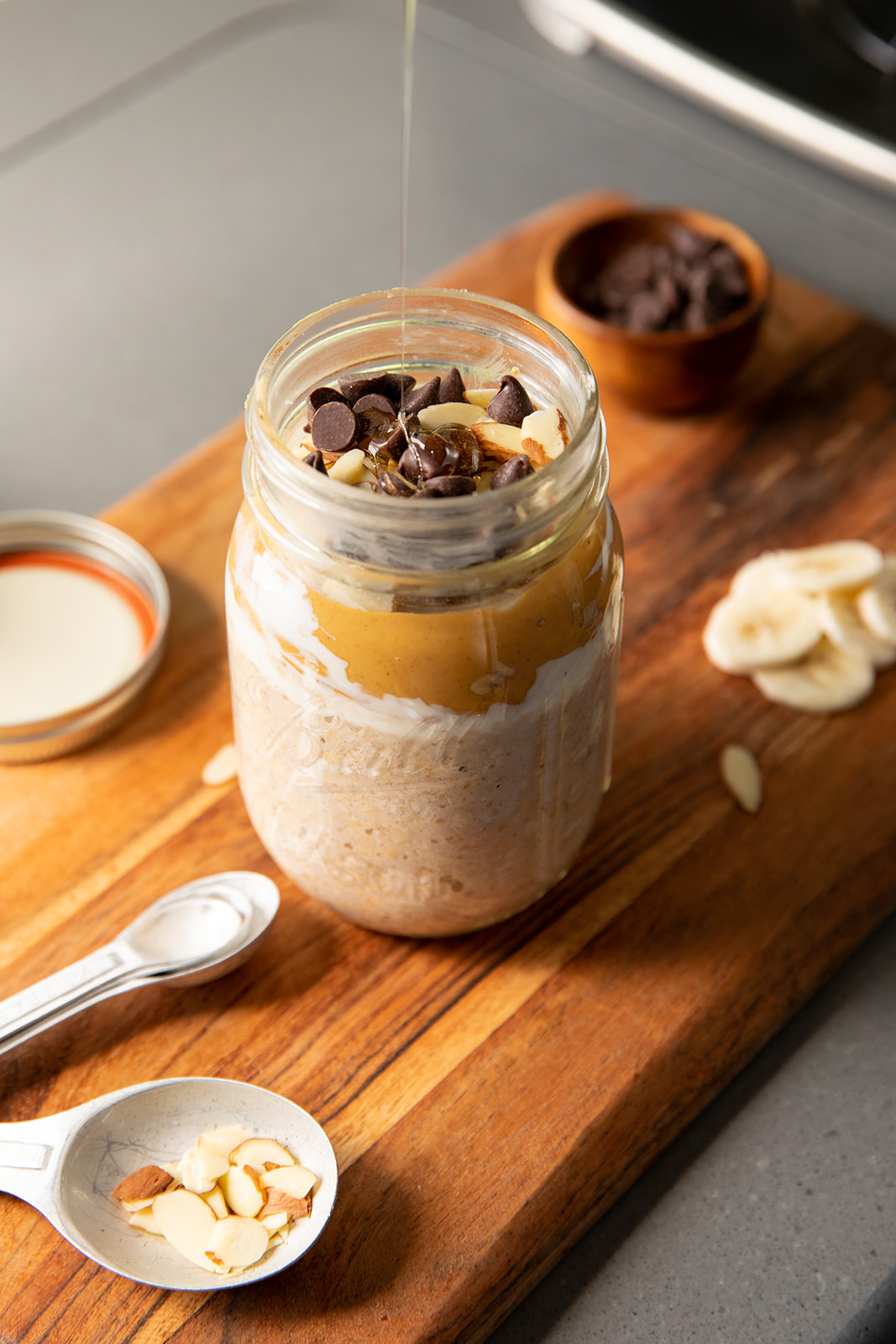 Honey drizzling into a mason jar with overnight oats topped with chocolate chips, peanut butter and almonds. 