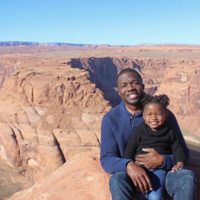 An African American father and daughter smile at the camera while sitting in front of Horseshoe Bend.