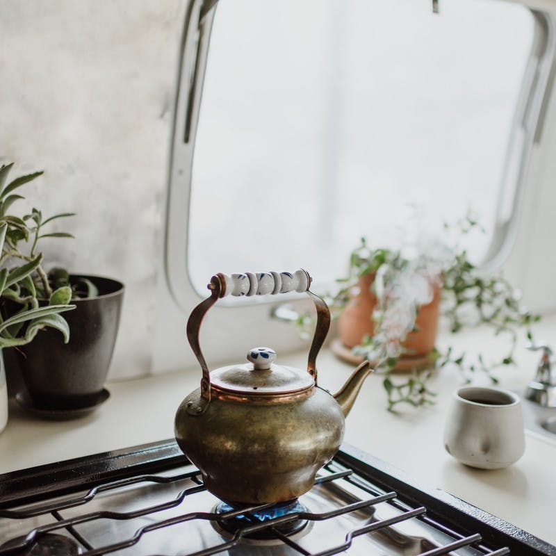 Close up of old copper tea kettle on a has stove, with plants and cups around it.