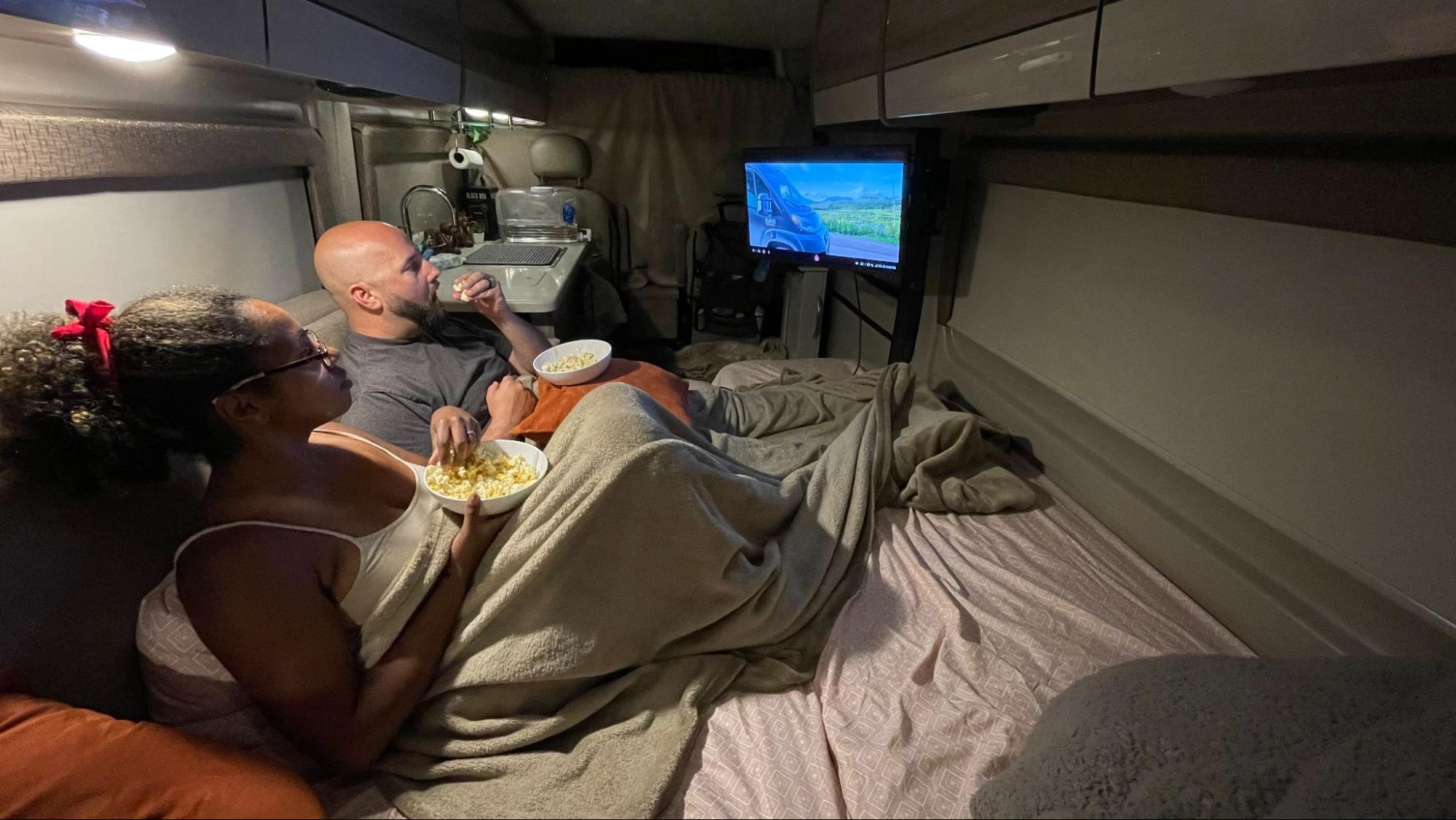 Rocio and Fabe eat popcorn and watching a movie while laying in the bed of their Class B RV.