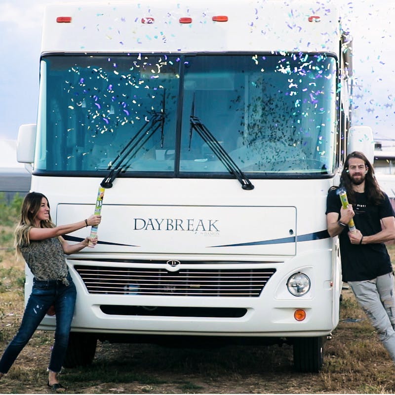 Bryce and Nellie Jurgy stand in front of the Class A motorhome, posing by windshield, popping streamers of colorful confetti into the air.
