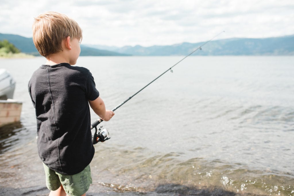 Young boy in black t-shirt holds fishing pole next to lake