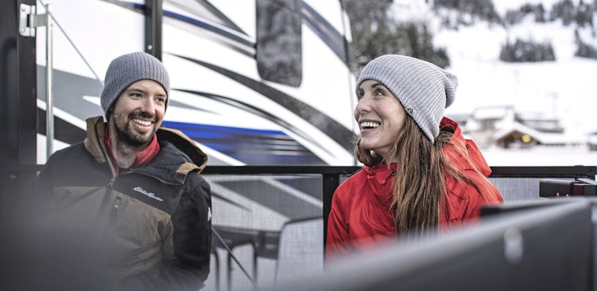 A young married couple smile together as they relax on the porch of a toy hauler in the mountains.