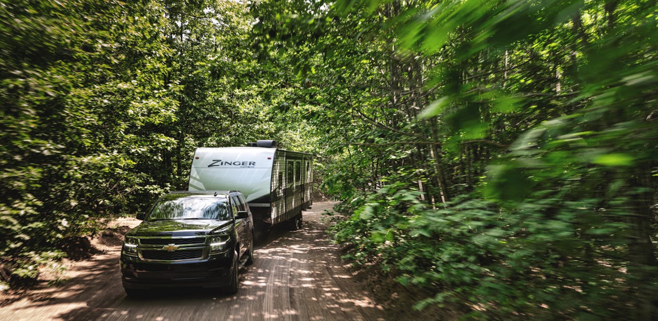 A truck tows a travel trailer down a wooded dirt road.