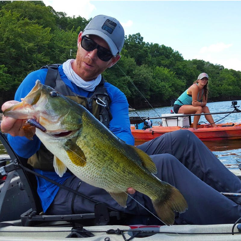 Robert holding up a bass from Lady Bird Lake as he sits in a kayak, with a friend in the background in another kayak.