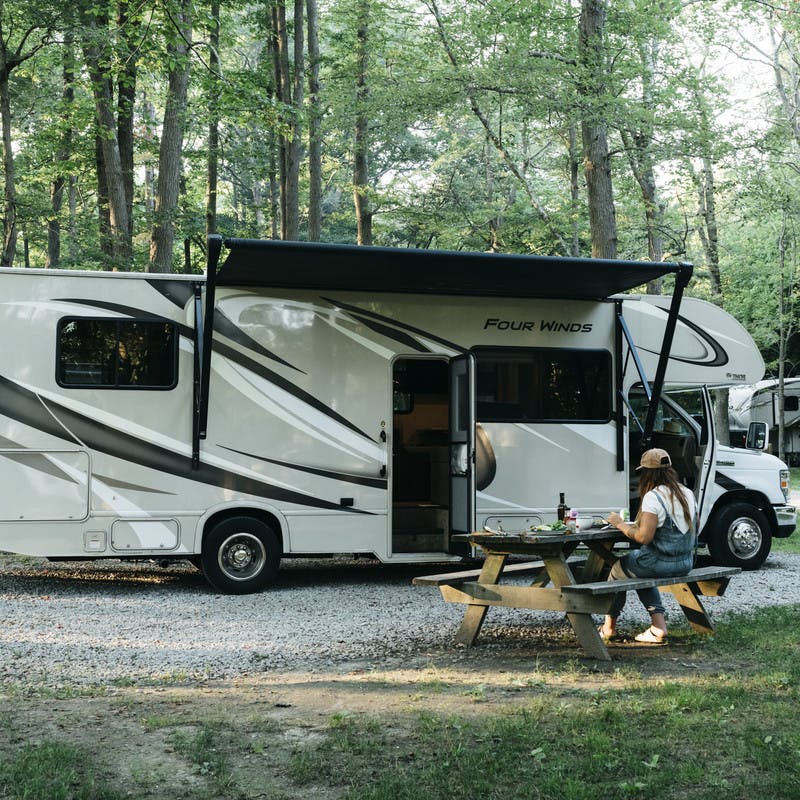 An RV at a tree-lined campground, parked with the awning up. Sarah sits at a picnic table nearby.
