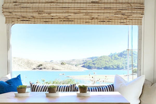 Interior shot of an RV, with a dining room table, chair, and bench filled with pillows for a comfortable seating area. The window behind the bench looks out to blue water and green trees on a sunny day.