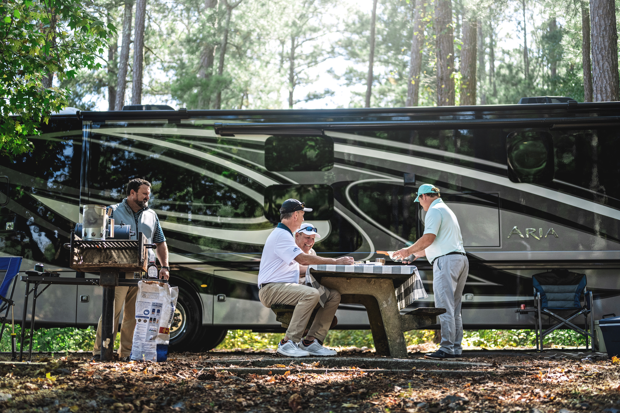 Men sit at a picnic table near an Aria Class A motorhome