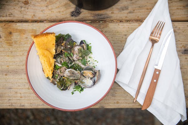 Bowl of cooked clams with parsley on a white plate, a golden slice of cornbread, and gold utensils on a white napkin.