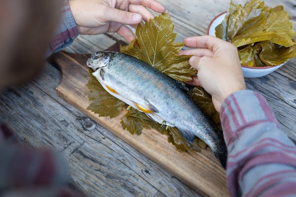 Hands wrapping full, stuffed trout in grape leaves, on a cutting board on a picnic table.
