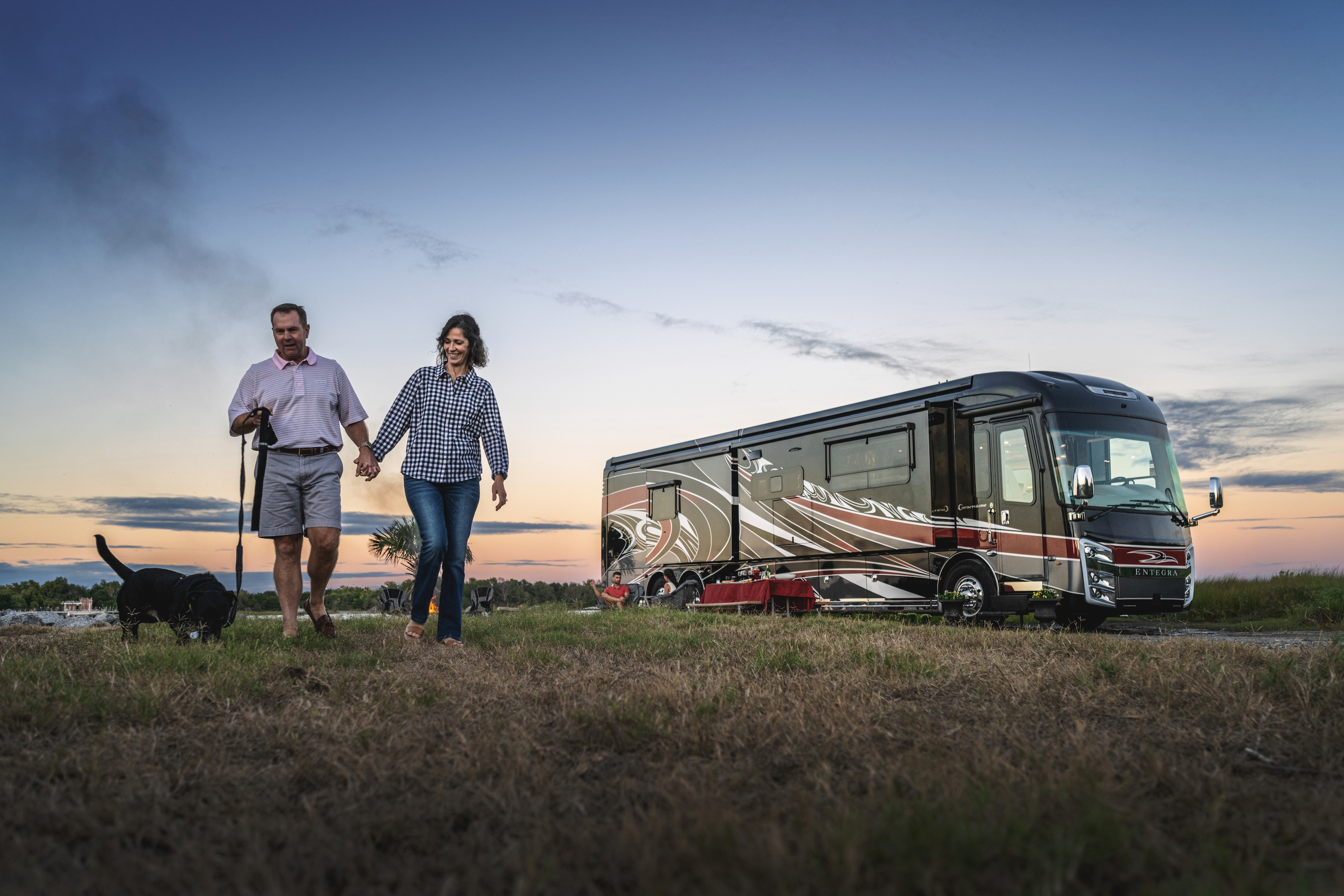 A couple walks a dog with a class A RV in the background as the sun sets.