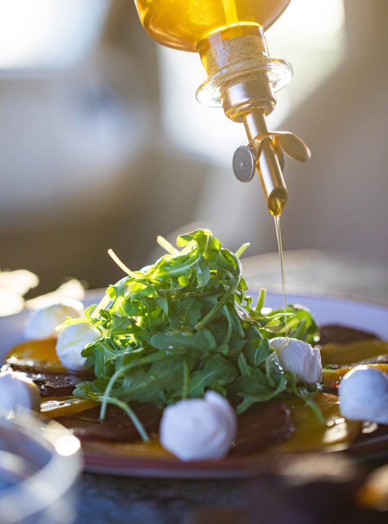Close up of olive oil being poured onto a pile of arugula lettuce and small mozzarella cheese balls.