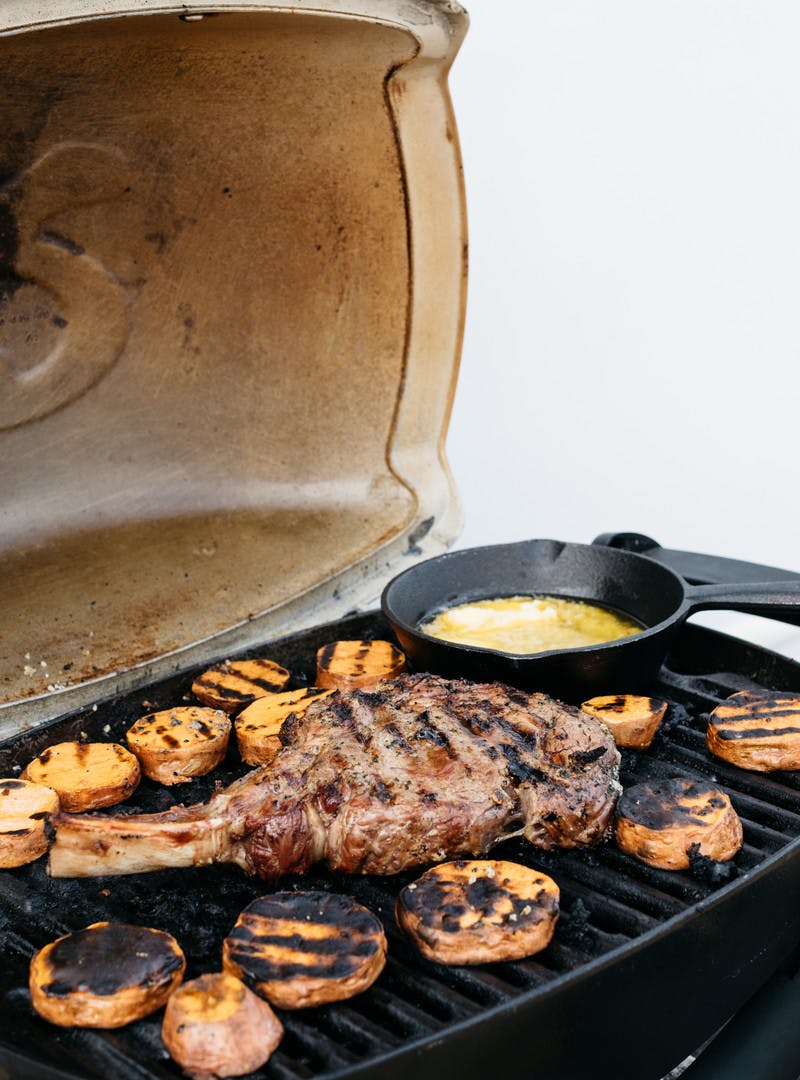 A steak with a long bone cooking on a grill alongside sweet potato rounds. 