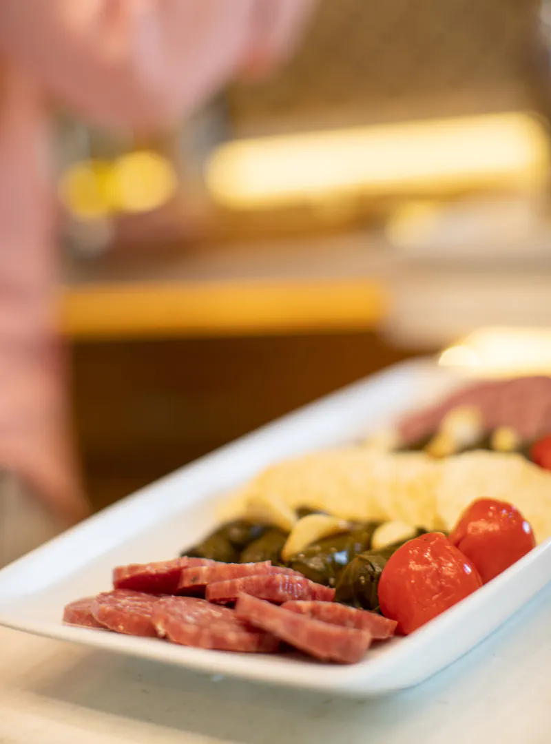 A white serving platter arranged with sliced salami, stuffed grape leaves, and red peppers.