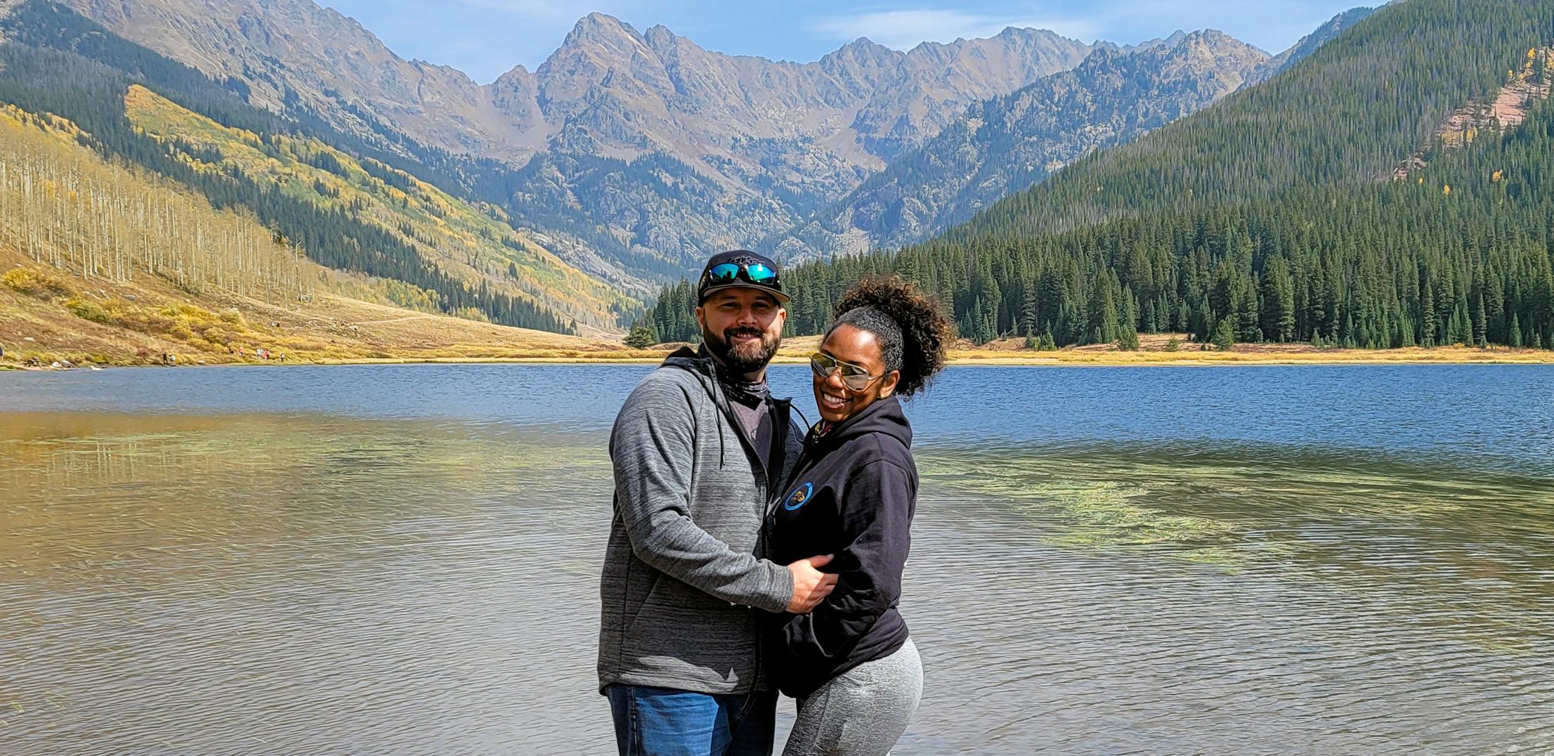 Gabe and Rocio Rivero pose at a scenic outlook featuring a lake, mountains, and forest.