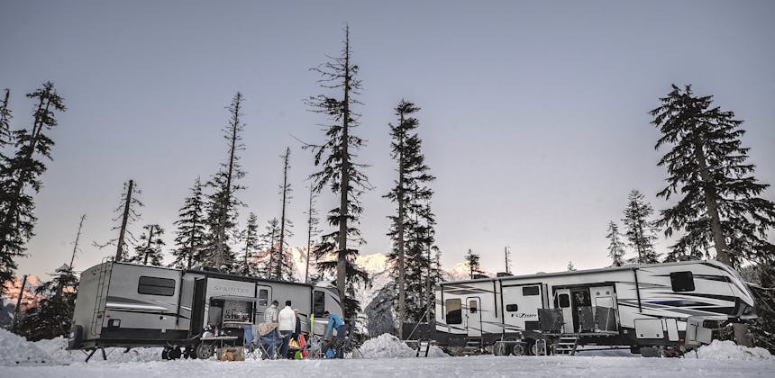 Two RVs stand side-by-side in the snowy mountains surrounded by trees.