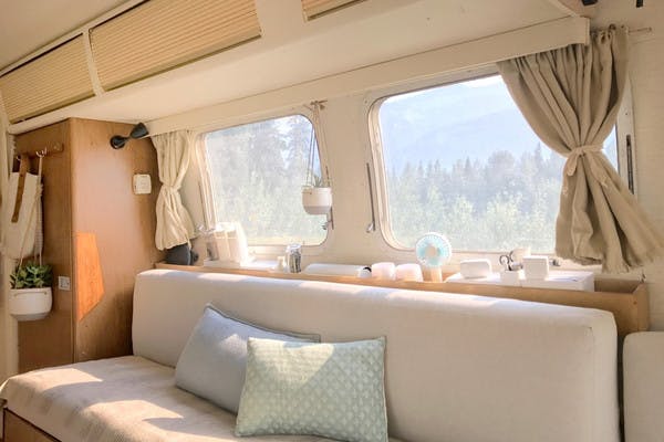 Interior shot of an RV, with cream colored couch, hanging plants and beige curtains, and pine trees outside the window.