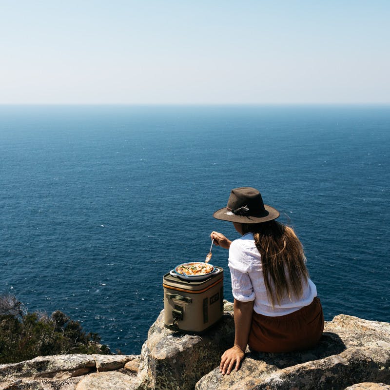 Sarah Glover sitting over the ocean eating cooked ricotta pasta shells from a pan.