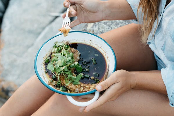 Close up of woman's legs, eating fish in brown broth with cilantro and a fork.
