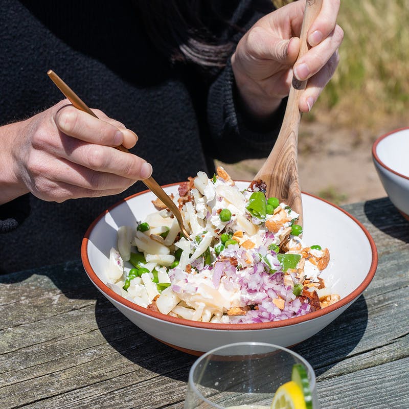 White bowl on picnic table mixing pea salad with wooden serving spoons.