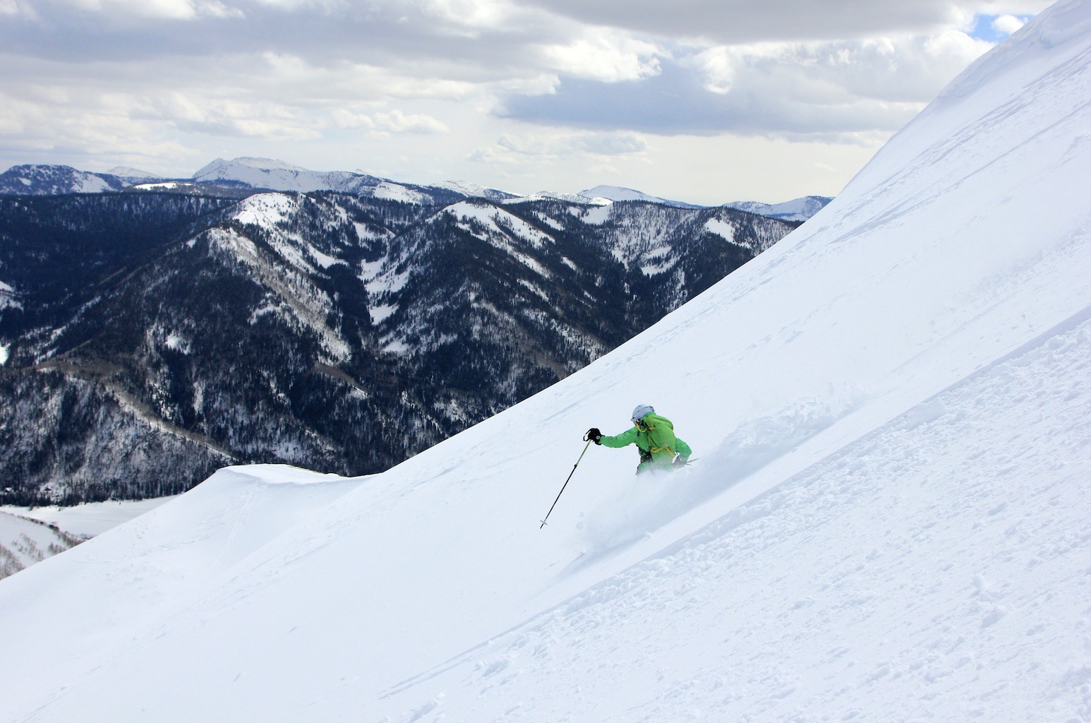 Man in green jacket skis down steep snowy mountain with dark mountains in the background