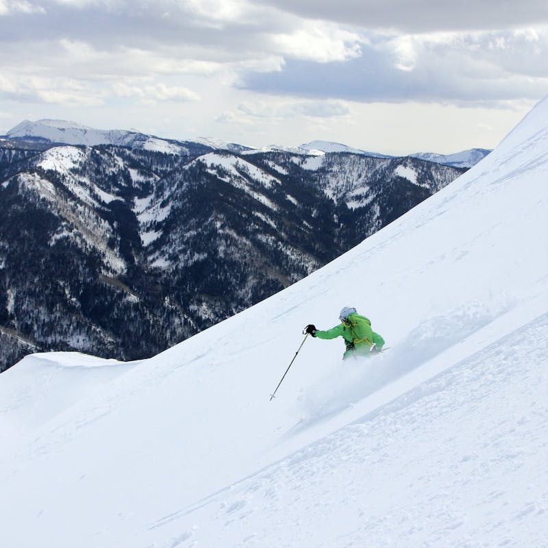 Man in green jacket skis down steep snowy mountain with dark mountains in the background