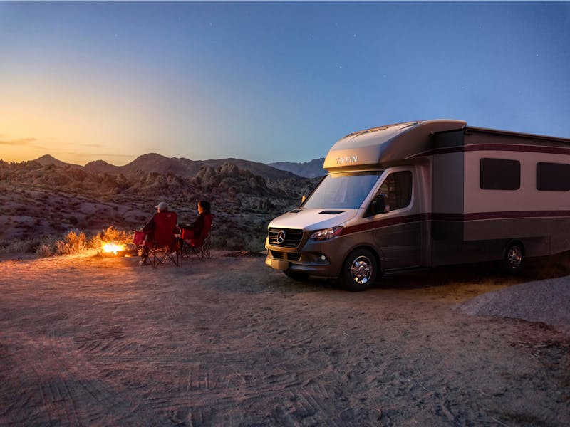 A Tiffin Class B Motorhome is parked in the desert with a couple enjoying a fire at dusk.