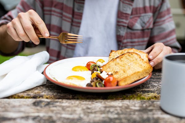 A plate of breakfast food, including eggs, toast, and vegetables, in front of a man holding a fork, about to break open the sunny-side eggs.