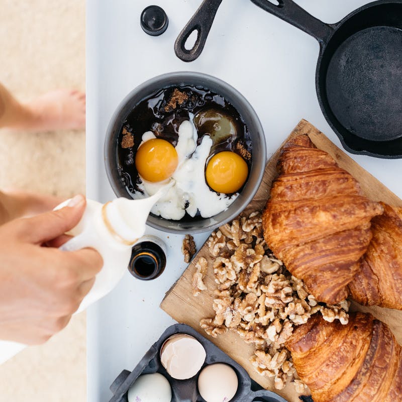 Top-down view of person standing at a counter, adding milk to a bowl holding cracked eggs and sugar. Croissants and chopped walnuts sit on a cutting board next to the bowl. 
