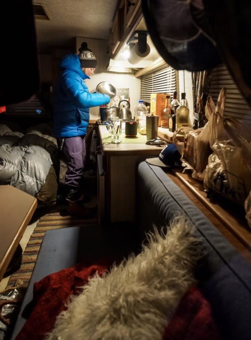 Blonde woman bundled up in a blue winter jacket, winter hat, winter pants, and boots standing in her RV kitchen cooking on the stovetop.