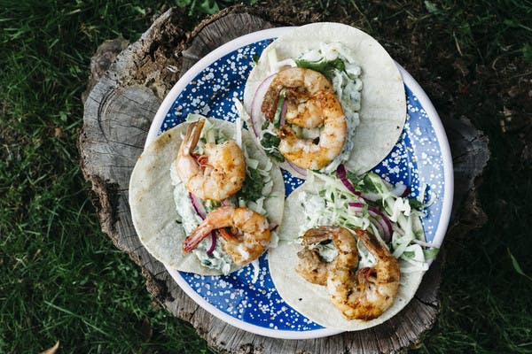 A plate of three assembled shrimp tacos, sitting on a tree stump outside.