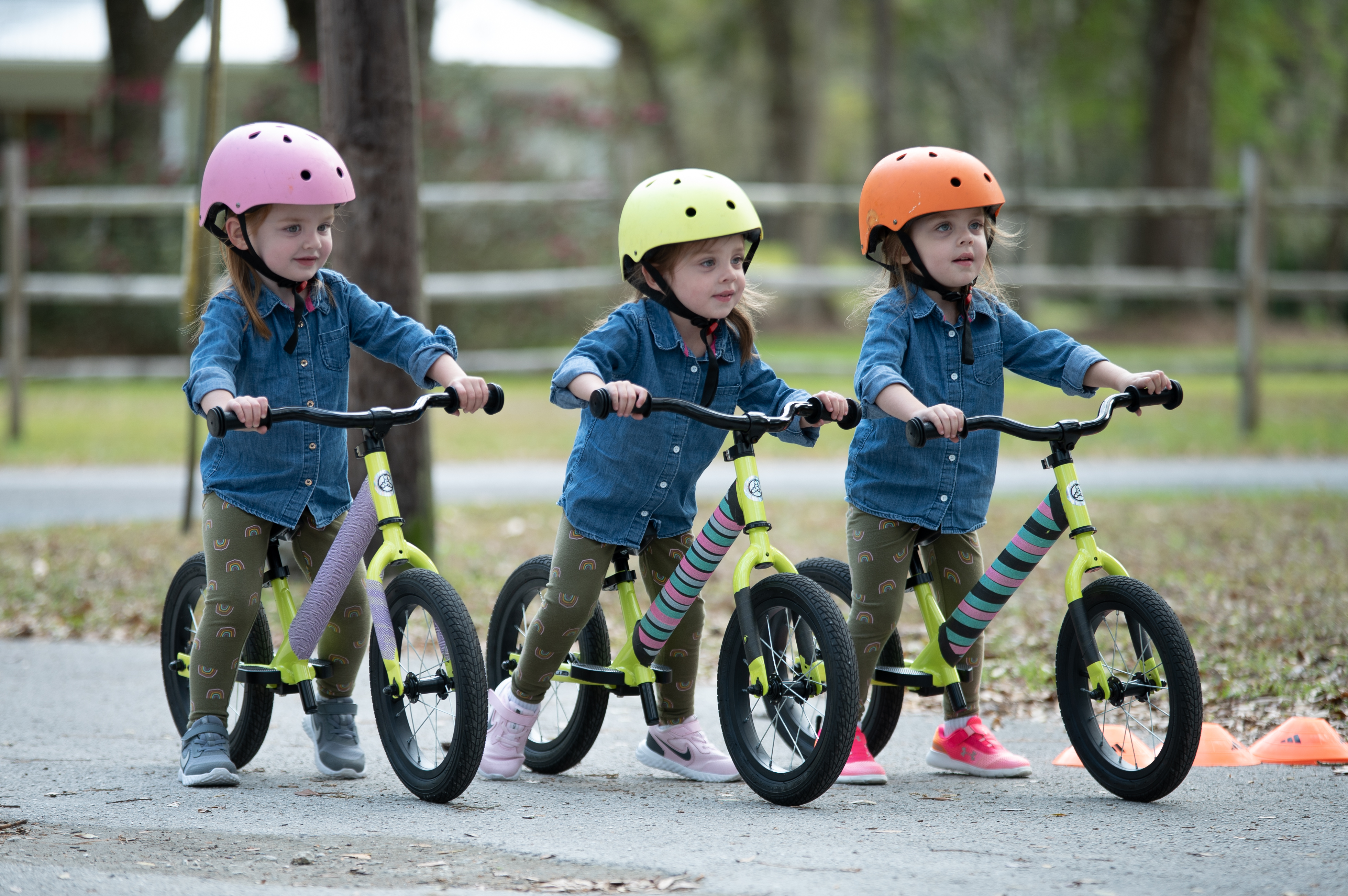 Triplet little girls riding bikes with color coded helmets on their heads.