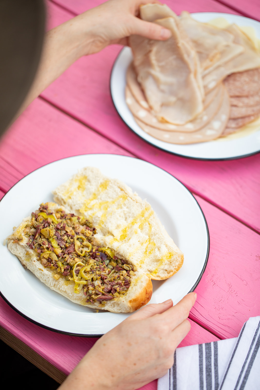 A hand grabbing lunchmeat to layer on a roll already dressed with olive tapenade.